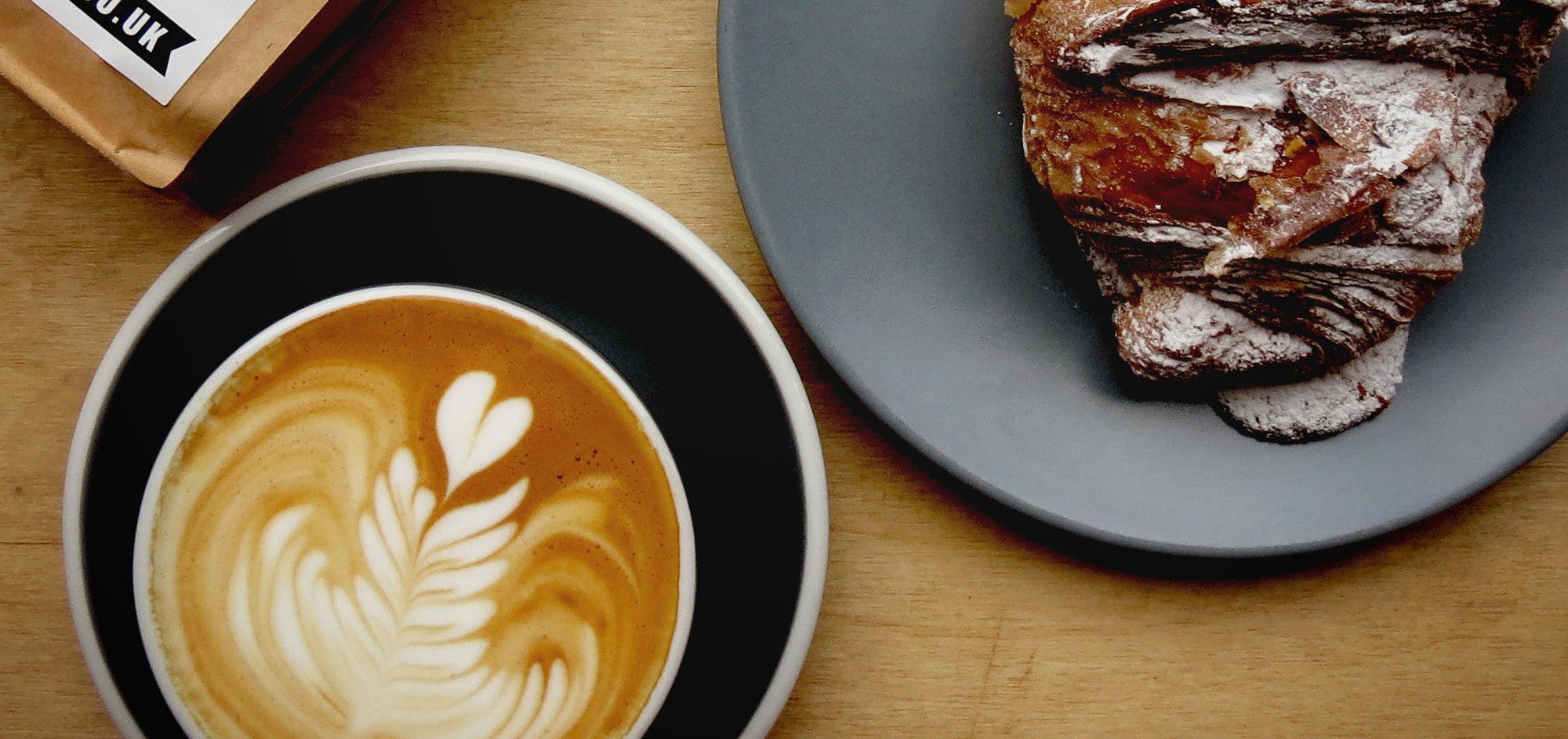 A flat white, a pastry and a bag of Coffee + Beer coffee on a wooden table